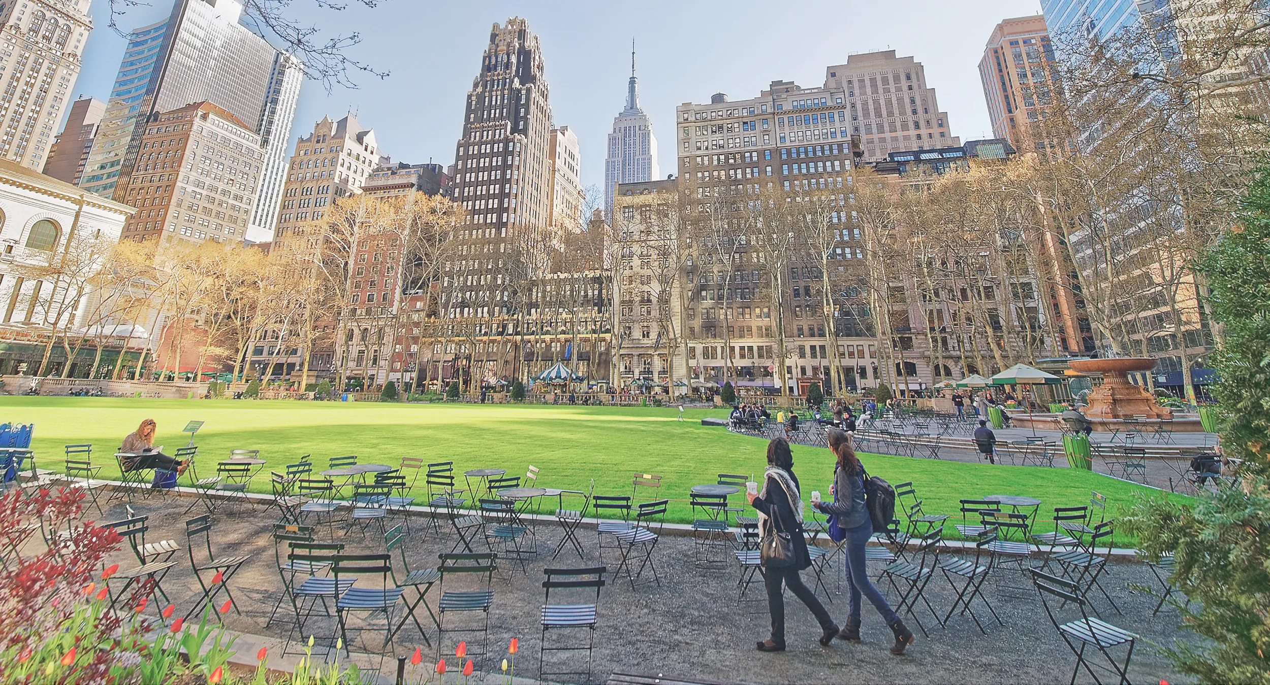 A photograph of a city park with high-rise buildings in the background. People sitting and walking among many chairs.